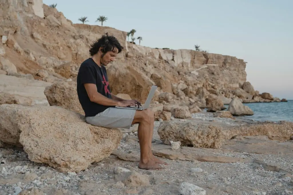 Man sits on rocks with laptop by the beach, enjoying remote Work in yucatán under a serene sunset.