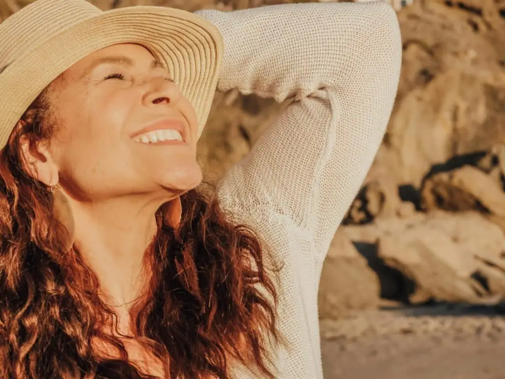 A joyful woman with a hat basks in the sunlight by the seaside, symbolizing relaxation and happiness. One Reason Expats Choose Yucatán.