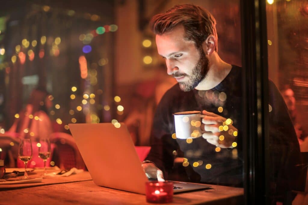 A young man with a beard is remote working in Yucatán on a laptop at a cozy cafe with warm lighting, holding a coffee cup.